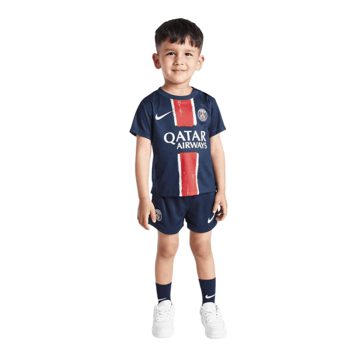 Smiling young boy standing in a navy PSG football kit with red vertical stripes and Qatar Airways logo, white Nike swoosh, matching shorts and socks against a white background.