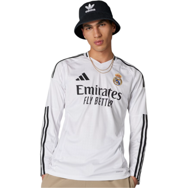 Young man wearing a white Real Madrid long-sleeve jersey with Emirates Fly Better logo and black Adidas stripes, paired with a black bucket hat and beige pants.