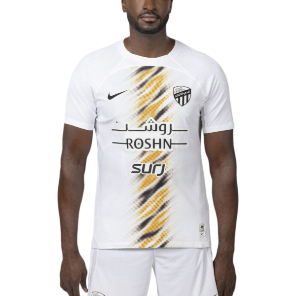 Man wearing a white soccer jersey with a yellow-black diagonal graphic and ROSHN sponsor logo on the chest.