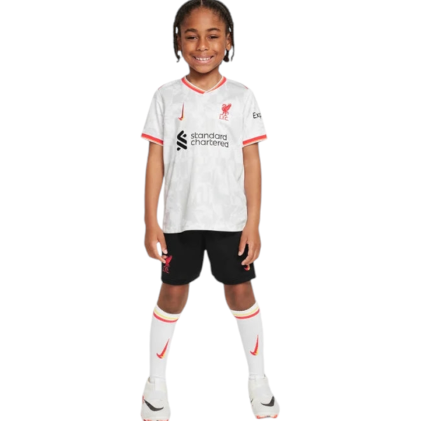 Child wearing a Liverpool FC home kit: white jersey with red trim, black shorts, white socks, standing and smiling.