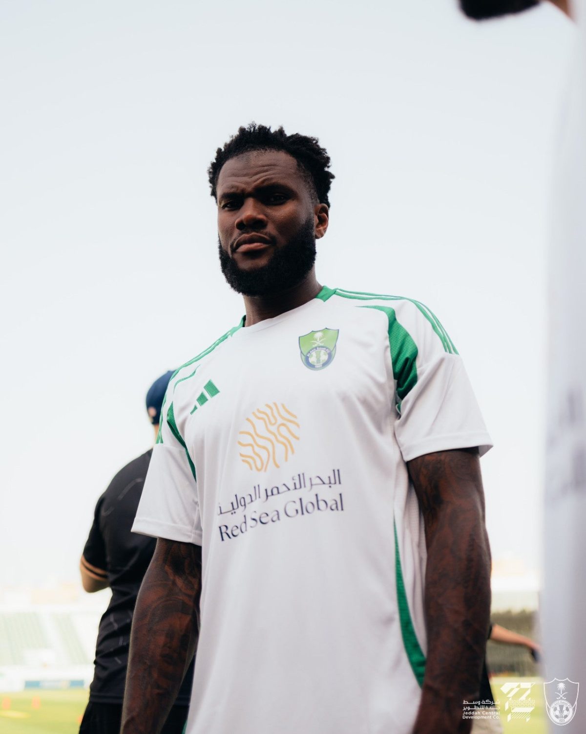 Black male soccer player wearing a white jersey with green accents and crest, standing on a field, looking toward the camera.
