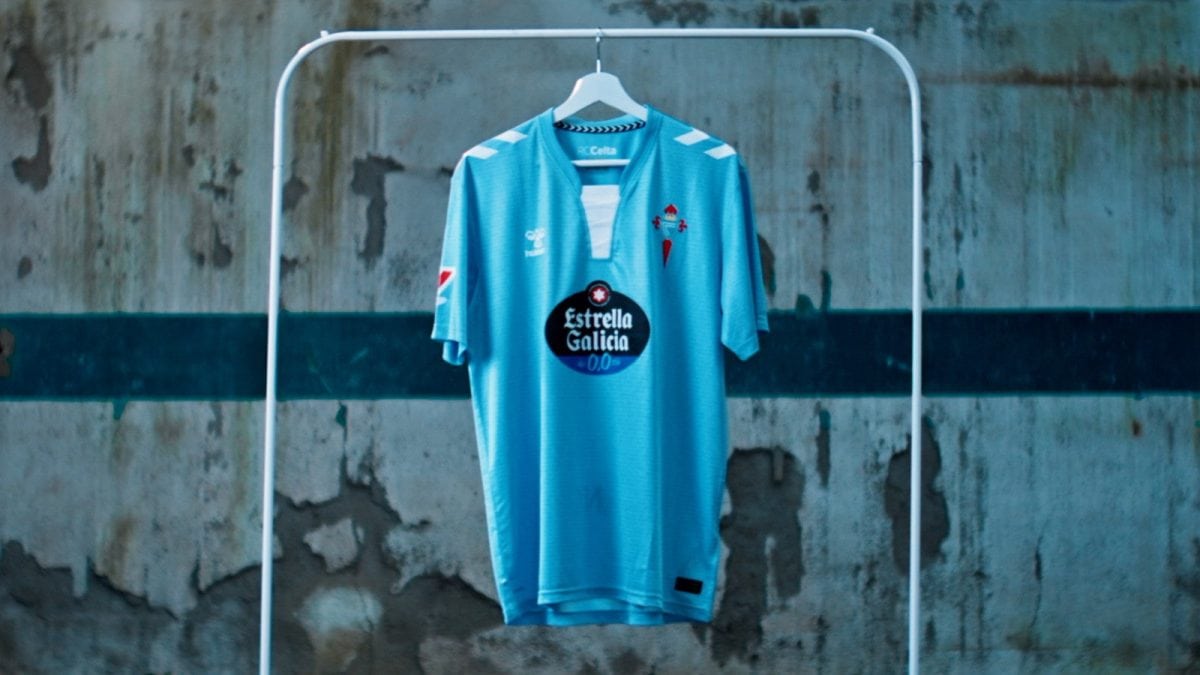 Blue soccer jersey on a white hanger inside a metal frame against a weathered wall; features Estrella Galicia sponsor logo on the chest and white chevrons on the shoulders.