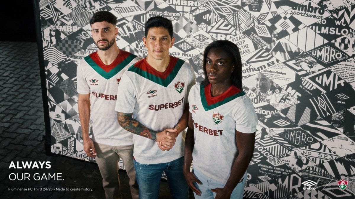 Three soccer players in white jerseys with red and green chevrons stand together; the center player shakes hands with a teammate while the others look at the camera against a patterned wall.