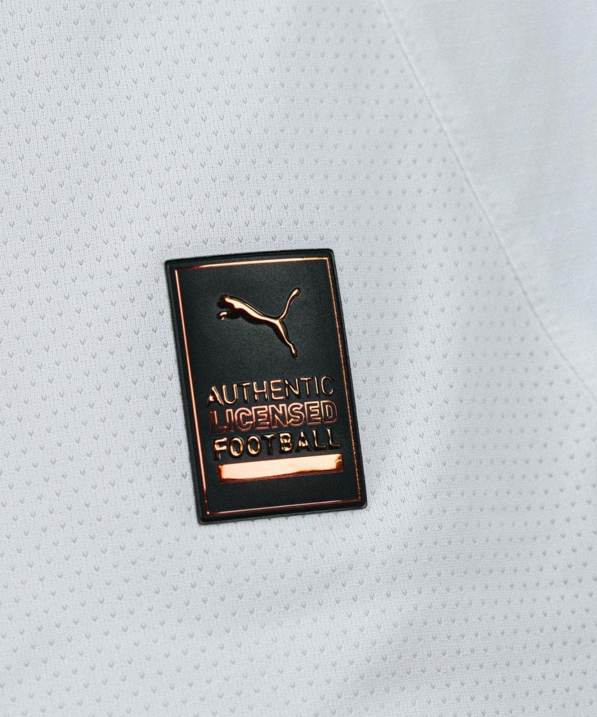 Metallic badge reading 'AUTHENTIC LICENSED FOOTBALL' with a Puma logo on a light grey perforated fabric background.