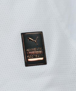 Metallic badge reading 'AUTHENTIC LICENSED FOOTBALL' with a Puma logo on a light grey perforated fabric background.