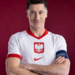 Portrait of a male footballer with arms crossed, wearing Poland's white jersey with the white eagle crest and a captain's armband against a red background.