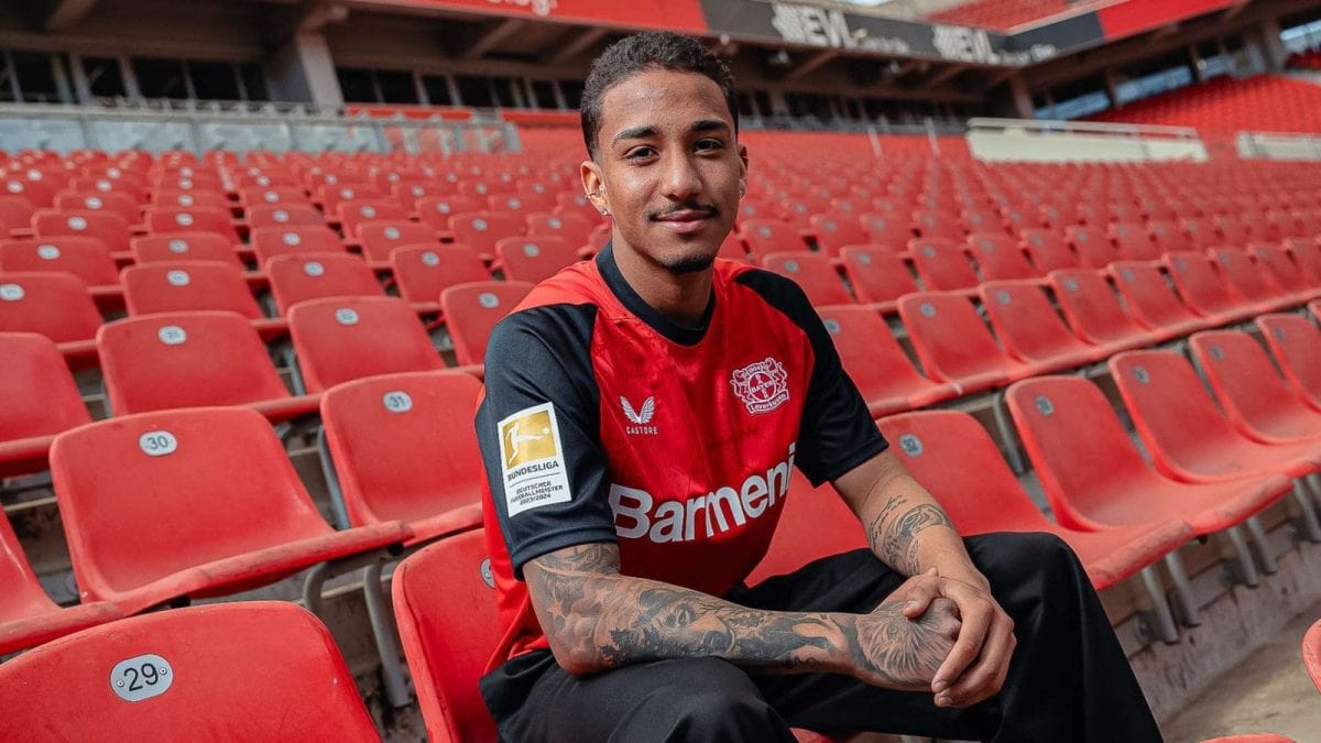 Portrait of a man in a red and black Bayer Leverkusen jersey sitting in empty red stadium seats, smiling.
