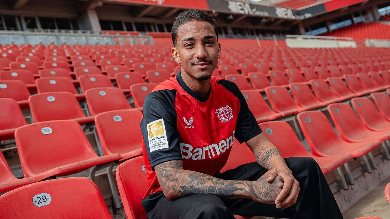 QUFrzb1pobuWYMz | UAE SPORT STORE Portrait of a man in a red and black Bayer Leverkusen jersey sitting in empty red stadium seats, smiling.