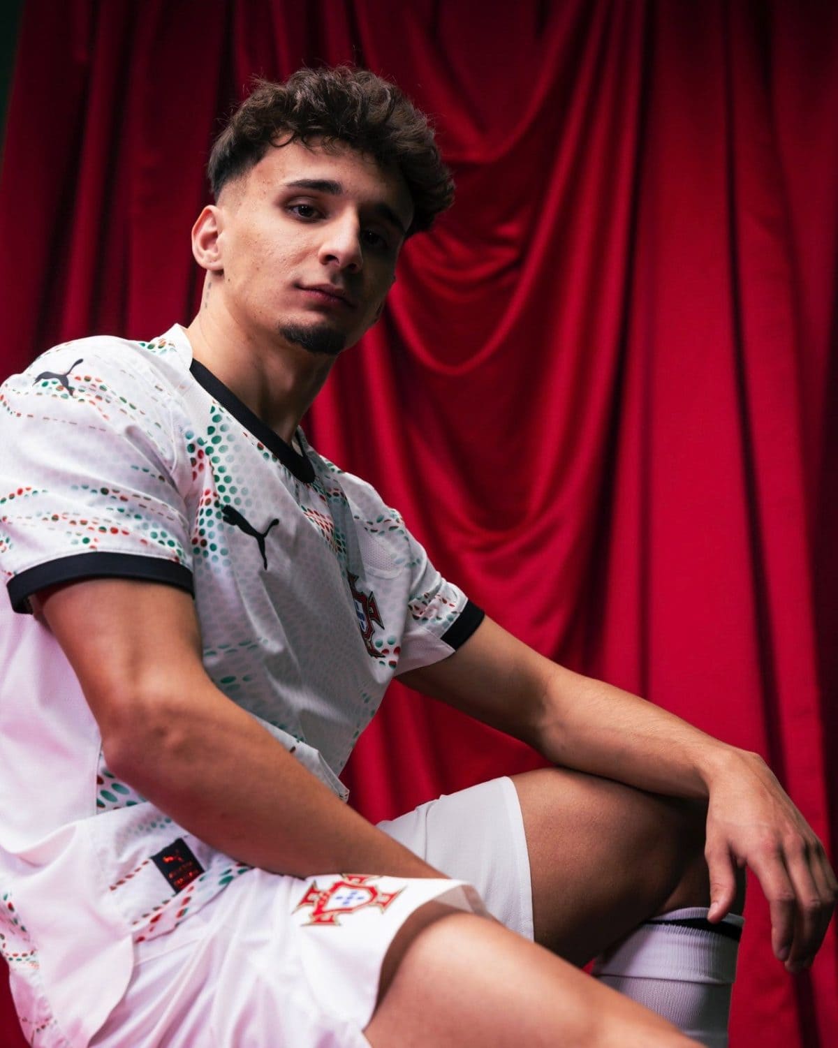 Young man in a white sports jersey with green and red patterns sits against a red curtain, looking at the camera.