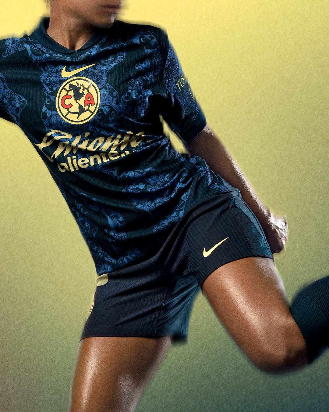 QVsY3zy1SvWxOXI | UAE SPORT STORE Athlete in a navy soccer jersey mid-kick, with gold crest and Nike swoosh visible against a yellow-green background into focus on the upper body and leg actions.
