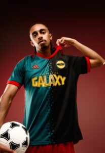 QYWWHY3HEqJChY6 | UAE SPORT STORE Portrait of a male soccer player wearing a teal and black Los Angeles Galaxy jersey, holding a soccer ball at his side.