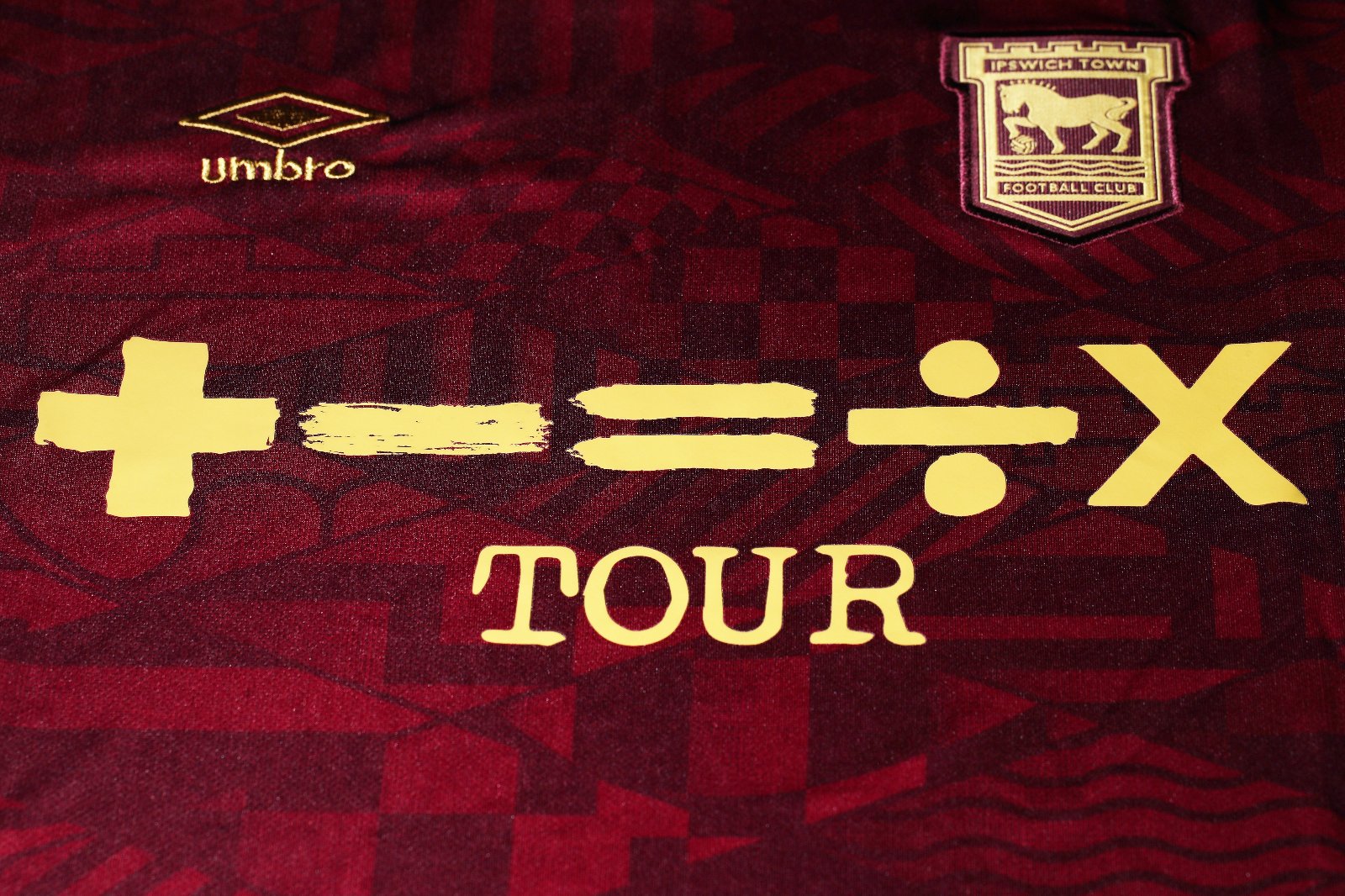 QlycHN8ymywpkzM | UAE SPORT STORE Close-up of a maroon Ipswich Town FC jersey with gold sponsor text and the club crest in the top right area, plus the Umbro logo near the left.