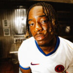 Close-up portrait of a young man with braided hair wearing a white jersey with blue trim indoors in a kitchen-like setting.