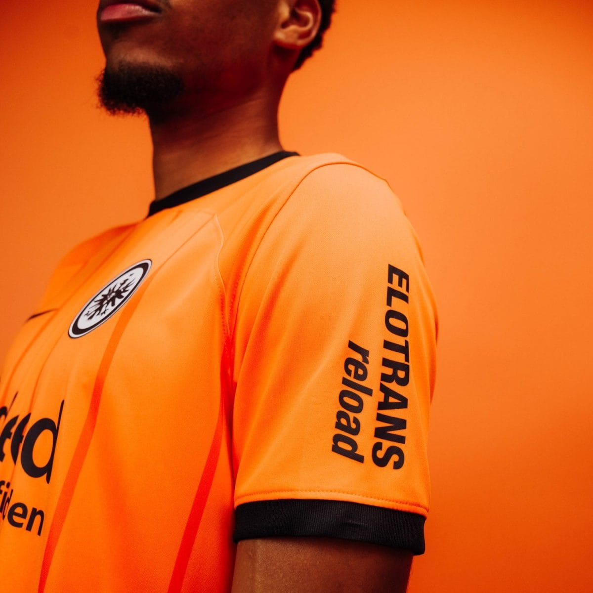 Close-up of a person in an orange athletic jersey with black trim, a circular chest logo, and black vertical text along the sleeve against an orange background.