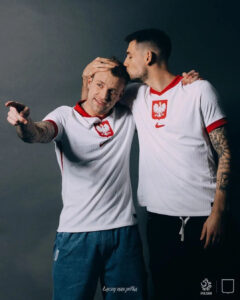 Two men in white Poland national team jerseys pose together; one kisses the other on the head while they smile and hug.