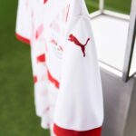 White sports jersey with red Puma logo on the sleeve, hanging on a rack near a grass field edge.