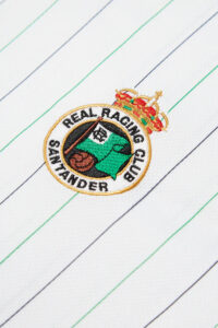 RACINGSHORTSLEEVE_03_600x_0792fe85-c0a2-47c9-99bc-f1cfb1df864c | UAE SPORT STORE Embroidered Real Racing Club crest on a white striped fabric with green and navy pinstripes.