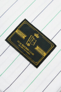 RACINGSHORTSLEEVE_06_600x_33c910ad-8bc9-47b5-8901-40d560edc4e6 | UAE SPORT STORE Black embroidered patch with gold border and insignia reading 'Real Racing Club' placed on white fabric with diagonal blue and green stripes.