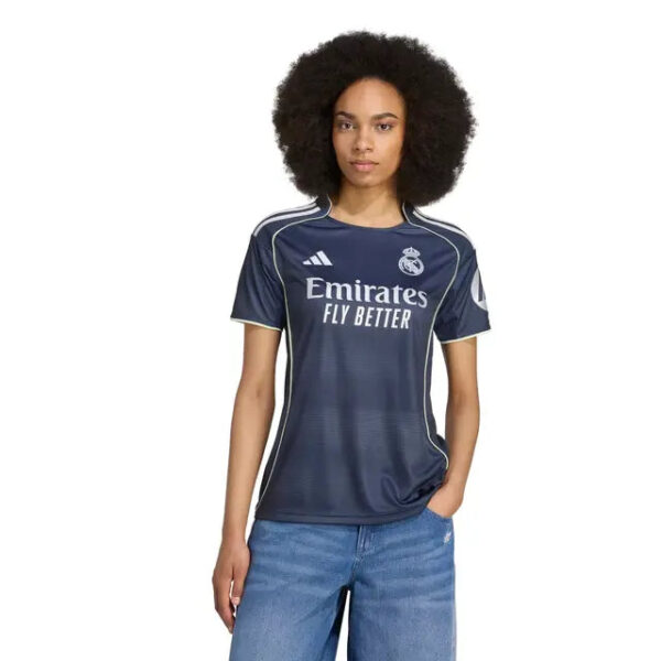 Woman wearing a navy Real Madrid jersey with Emirates Fly Better logo, standing with one hand in her pocket outdoors (product shot).