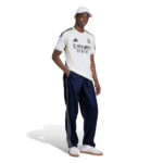 Male model wearing Real Madrid white jersey with Emirates Fly Better text, navy track pants, white cap, and sneakers posing against a white background.