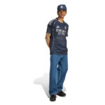 Man modeling a Real Madrid navy away kit: cap, matching jersey with Emirates Fly Better logo, and loose blue jeans with black sneakers.