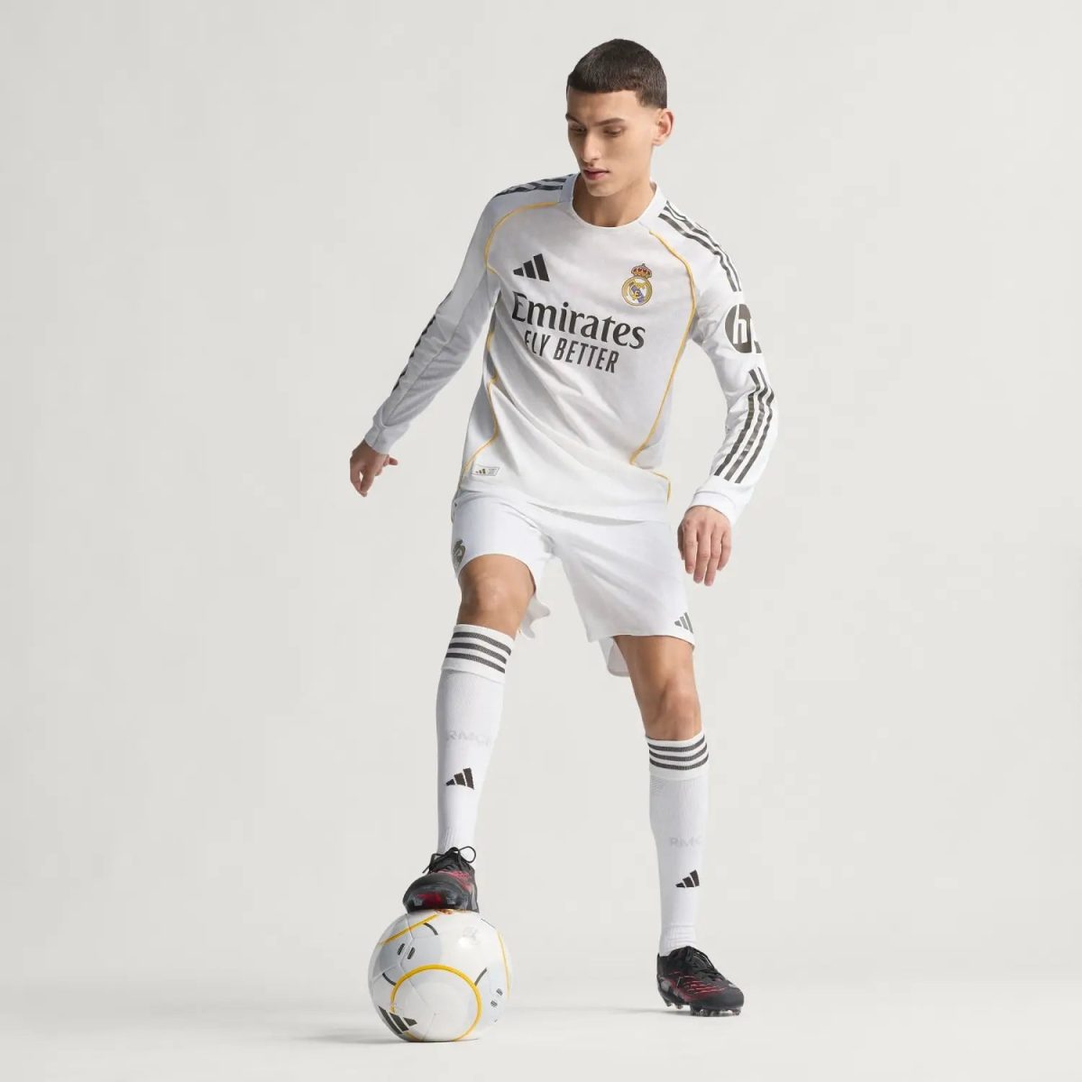 Male soccer player in a Real Madrid white kit balancing a ball with his foot, posed in a neutral studio background (advertisement-style image).