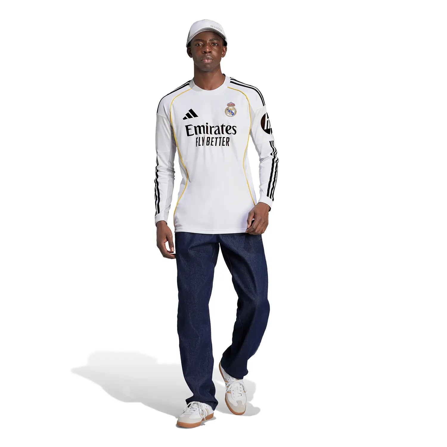 RMCFMZ0905_05 | UAE SPORT STORE Male model wearing Real Madrid white long-sleeve jersey with Emirates Fly Better sponsor, black Adidas stripes, and yellow piping, paired with dark jeans and white sneakers.