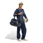 Athlete in Real Madrid training gear holding a gym bag over the shoulder and a soccer ball under the arm, wearing a cap and sneakers.
