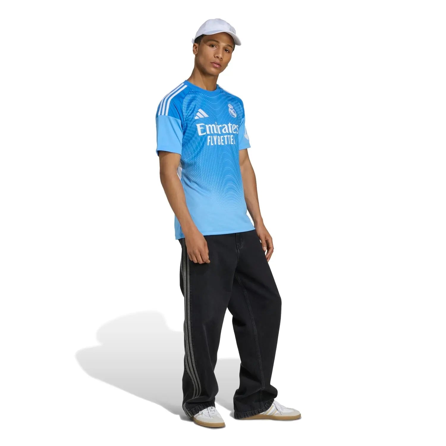 RMCFMZ0916_06 | UAE SPORT STORE Male model wearing a light blue Real Madrid jersey with Emirates Fly Better sponsor, white cap, and black pants with white sneakers.