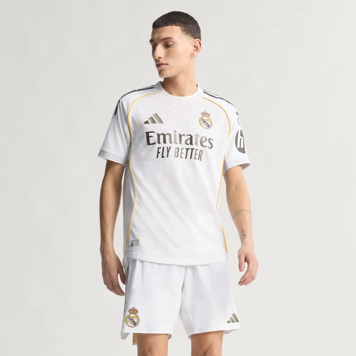 Man wearing Real Madrid white soccer jersey and shorts, standing against a light background; jersey features Emirates Fly Better logo and Real Madrid crest on chest, with yellow piping along seams.