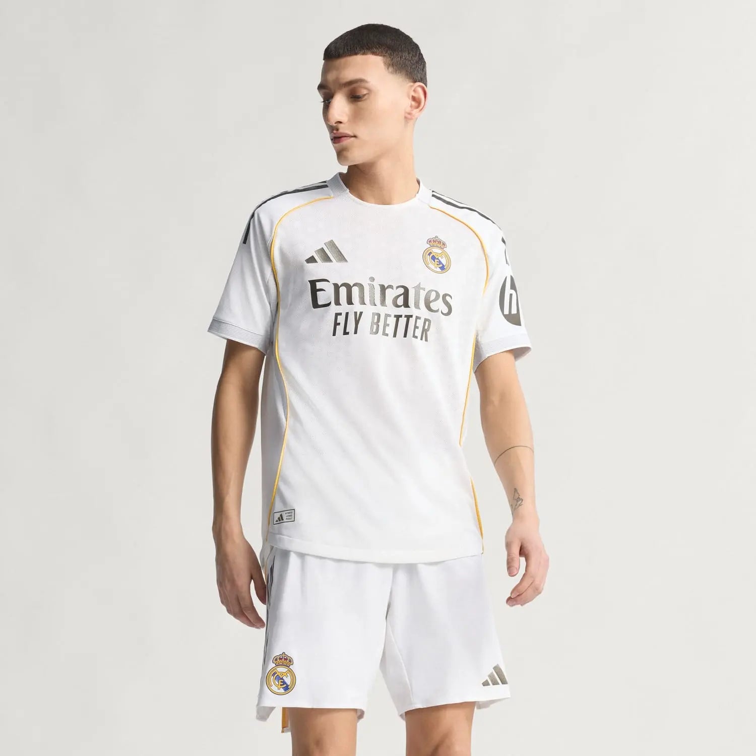 RMCFMZ0919_05 | UAE SPORT STORE Man wearing Real Madrid white soccer jersey and shorts, standing against a light background; jersey features Emirates Fly Better logo and Real Madrid crest on chest, with yellow piping along seams.