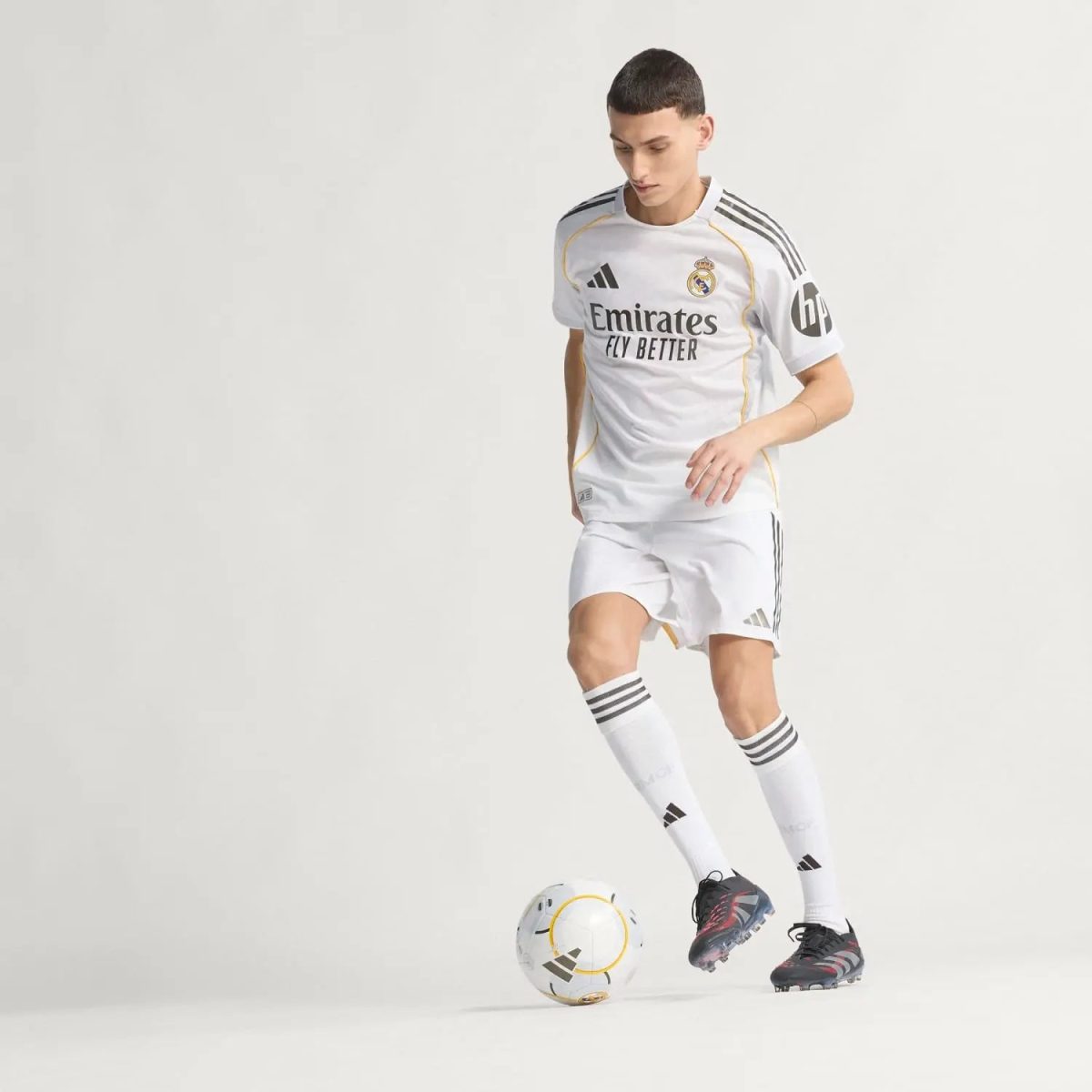 Soccer player in a white Real Madrid kit dribbling a ball on a plain light background, showcasing Emirates Fly Better sponsor.