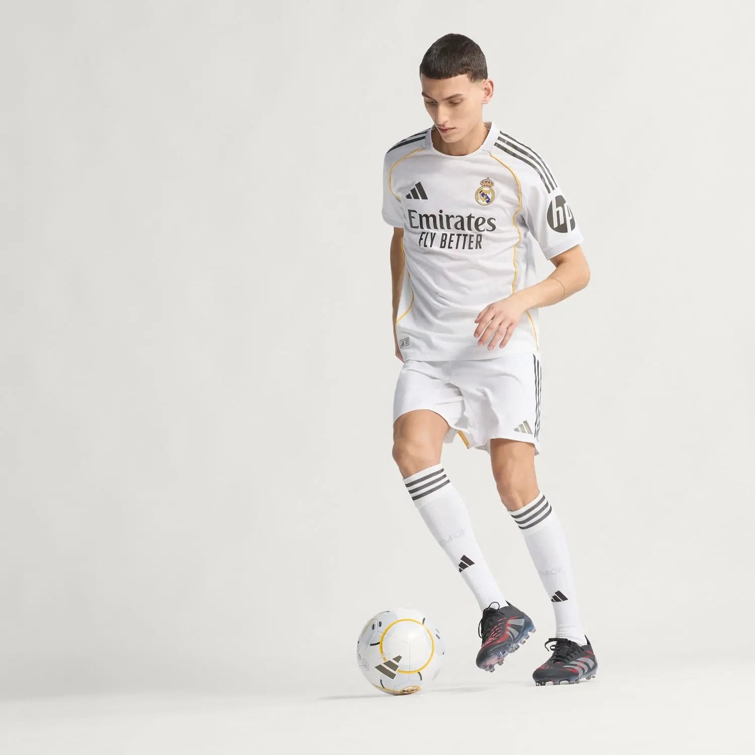 RMCFMZ0919_07 | UAE SPORT STORE Soccer player in a white Real Madrid kit dribbling a ball on a plain light background, showcasing Emirates Fly Better sponsor.