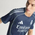 RMCFMZ0920_13 | UAE SPORT STORE Person wearing a navy Real Madrid soccer jersey with white shoulder stripes and ‘Emirates Fly Better’ text, shown from the chest up.