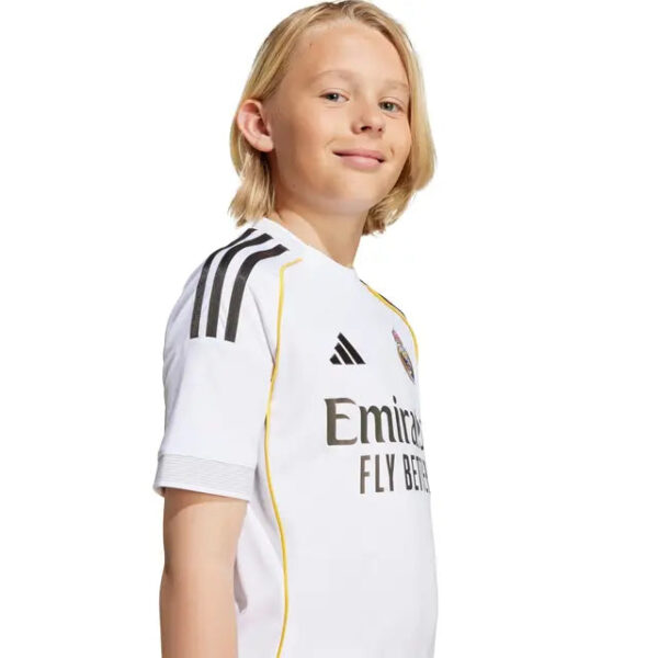 Young boy wearing a white Real Madrid jersey, side profile, smiling slightly at the camera, against a white background.