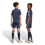 Two children in matching navy sports uniforms stand back-to-back; the girl faces slightly toward the camera with a smile while the boy faces away. They wear navy jerseys, shorts, long socks, and light-colored sneakers.