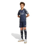 RMCFYO0095_6 | UAE SPORT STORE Boy wearing a dark Real Madrid soccer jersey with matching shorts and long socks, standing with hands behind back and white sneakers.