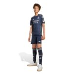 Young boy standing in a navy Real Madrid kids football kit, jersey and shorts, with matching socks and sneakers, sponsor logo visible.