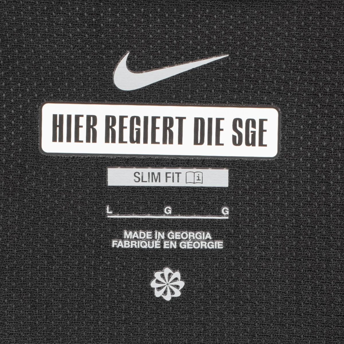 Nike logo above a white label reading 'HIER REGIERT DIE SGE' on a black textured surface, with 'SLIM FIT' and a small icon beneath and 'MADE IN GEORGIA' text further down.