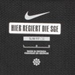 RNw4dH1UIzsoGJx | UAE SPORT STORE Nike logo above a white label reading 'HIER REGIERT DIE SGE' on a black textured surface, with 'SLIM FIT' and a small icon beneath and 'MADE IN GEORGIA' text further down.