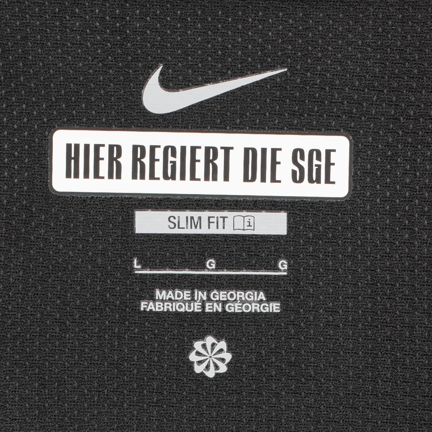 RNw4dH1UIzsoGJx | UAE SPORT STORE Nike logo above a white label reading 'HIER REGIERT DIE SGE' on a black textured surface, with 'SLIM FIT' and a small icon beneath and 'MADE IN GEORGIA' text further down.