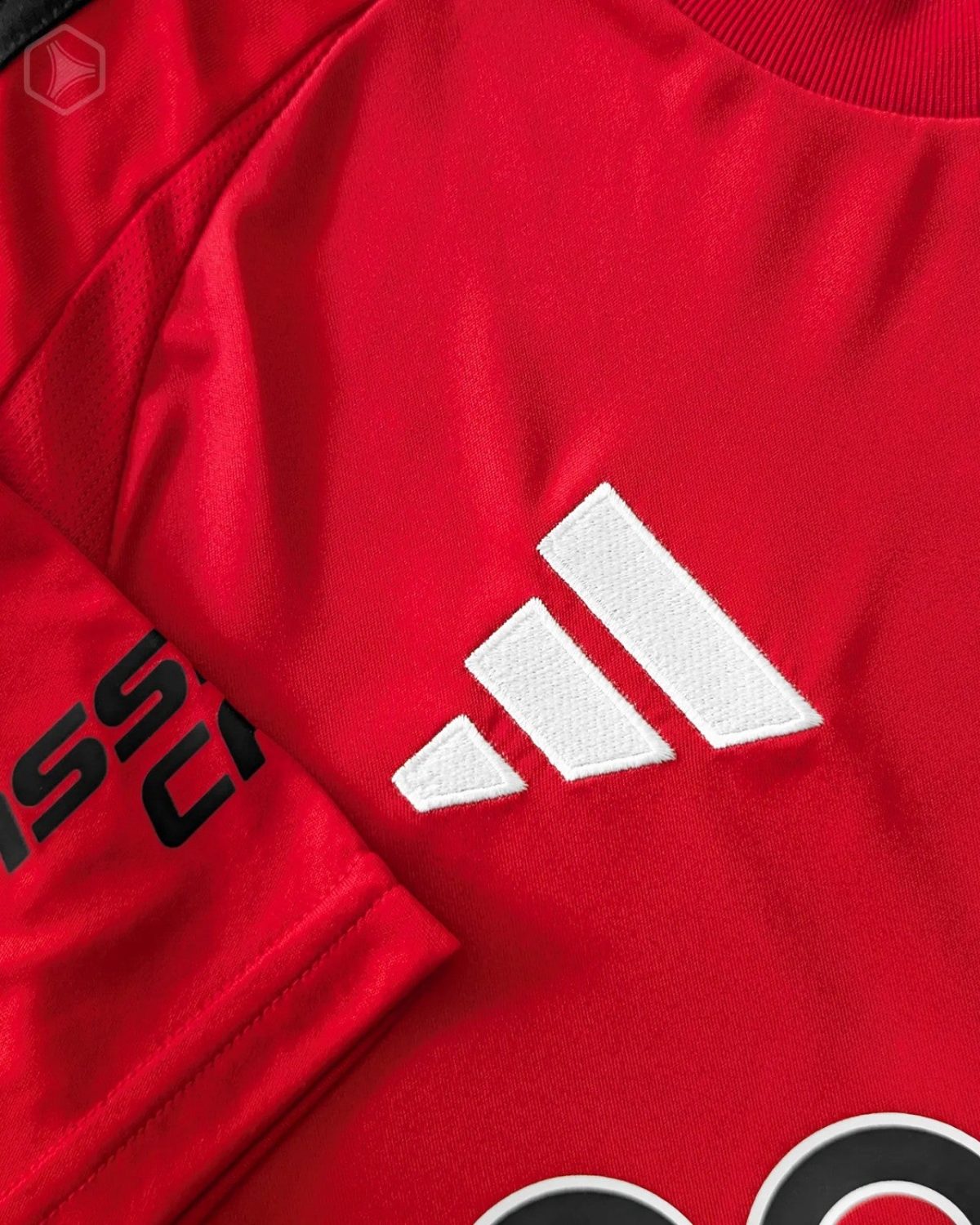 Close-up of a red Adidas jersey showing the white embroidered trefoil/three-stripe logo on fabric.