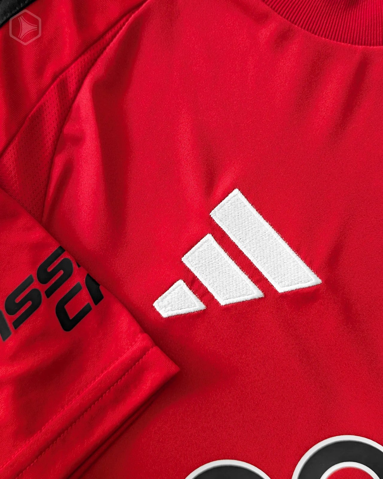 RZJnJEdVbD6tTQy | UAE SPORT STORE Close-up of a red Adidas jersey showing the white embroidered trefoil/three-stripe logo on fabric.