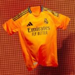Rdwqv1eoxejVNVD | UAE SPORT STORE Bright orange Real Madrid football jersey with Emirates Fly Better sponsor, club crest, and Adidas stripes on the shoulders, displayed against a matching orange background.