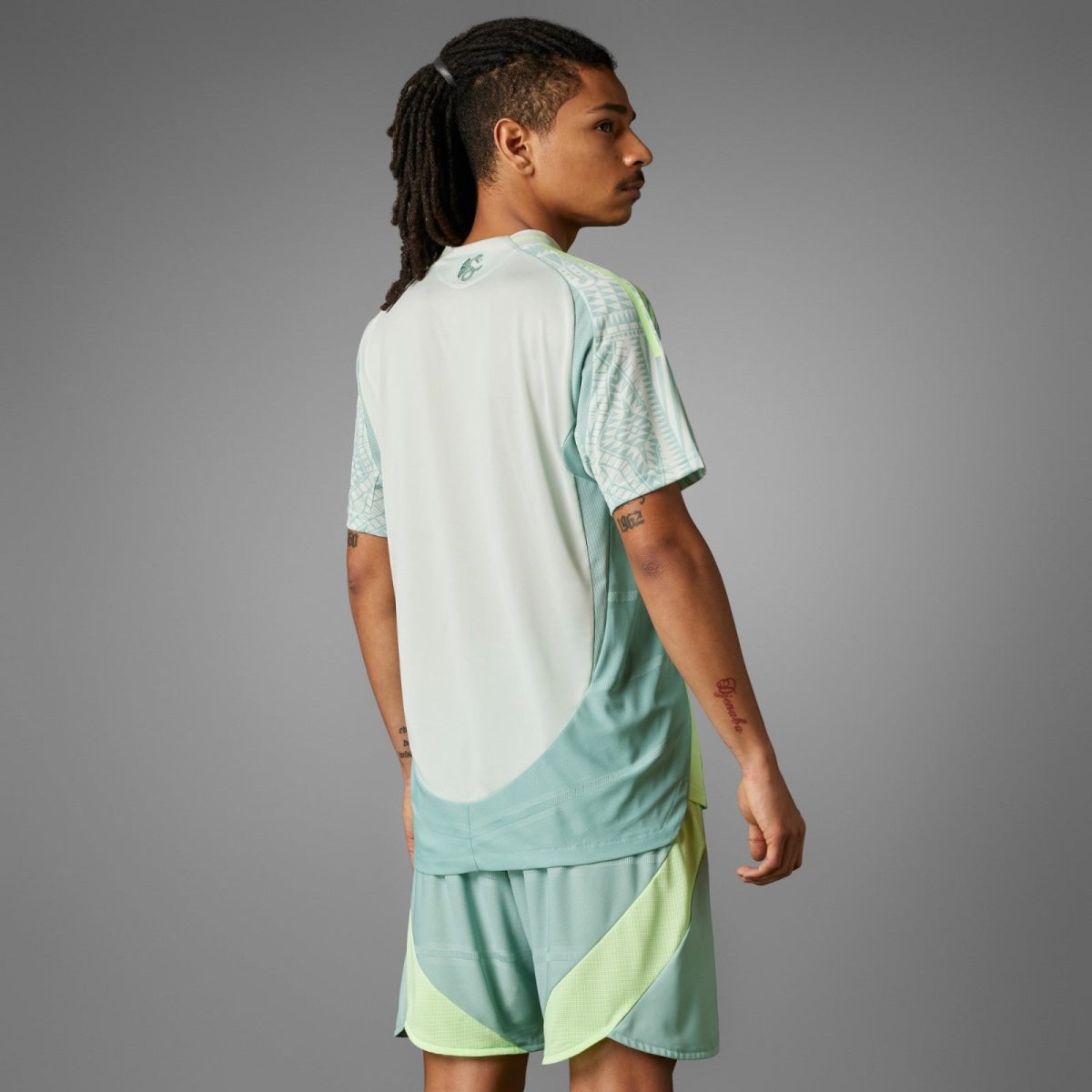 Side view of a young man with dreadlocks wearing mint green athletic jersey with geometric sleeve pattern, seen from behind against a gray backdrop.