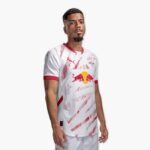 Male soccer player modeling a white RB Leipzig jersey with red accents against a plain light background, displaying sponsor logos (Red Bull) on the chest.