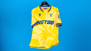 Yellow soccer jersey on a hanger against a solid blue background, featuring a blue NET88 sponsor logo and club crest.