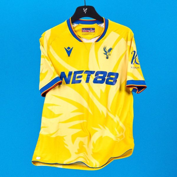 Yellow soccer jersey on a hanger against a solid blue background, featuring a blue NET88 sponsor logo and club crest.
