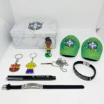 A collection of soccer-themed memorabilia on a white surface: a marble-patterned metal tin, a small Brasil/CBF figurine, two green Brazil-branded items, keychains, a necklace, a black bracelet, a pen, and a watch strap.