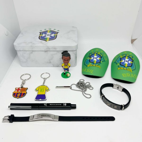 A collection of soccer-themed memorabilia on a white surface: a marble-patterned metal tin, a small Brasil/CBF figurine, two green Brazil-branded items, keychains, a necklace, a black bracelet, a pen, and a watch strap.